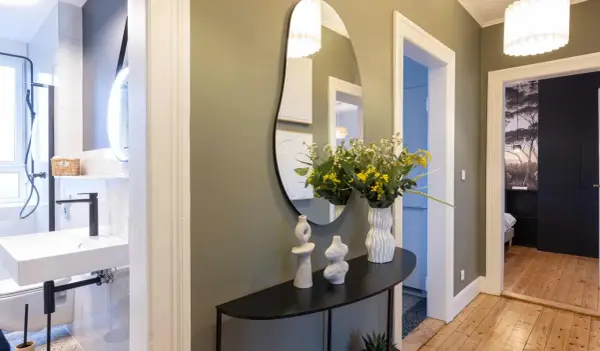 Élégant couloir maison avec miroir asymétrique, vases décoratifs, bouquet de fleurs jaunes et vue sur une salle de bain et une chambre.